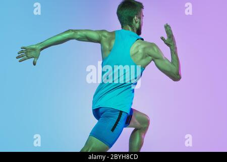 Portrait of active young caucasian man running, jogging on gradient studio background in neon light. Professional sportsman training in action and motion. Sport, wellness, activity, vitality concept. Stock Photo