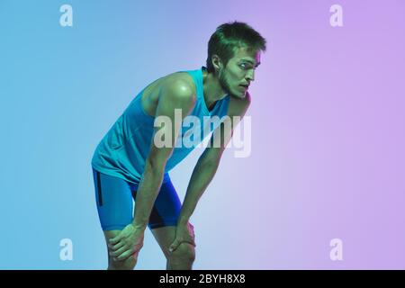 Resting. Portrait of young caucasian man running, jogging on gradient studio background in neon light. Professional sportsman training in action and motion. Sport, wellness, activity, vitality concept. Stock Photo