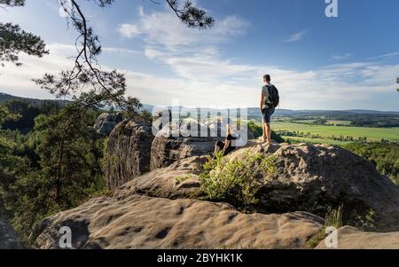 Hikers with backpacks relaxing on top of a mountain and enjoying the view of valley on the edge sandstone rock towers beautiful landscape in Bohemian Stock Photo