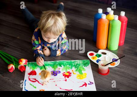 Sweet toddler blond child, boy, painting with colors, little chicks walking around him, making funny prints on his paper Stock Photo
