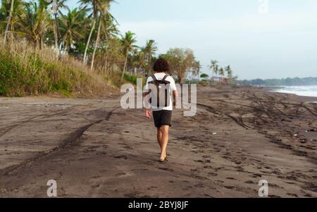 Rear view of fashionable male with curly haired student wearing brown leather backpack on black seashore alone, facing azure sea, came palm beach. Stock Photo