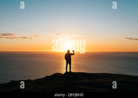Silhouette of a young man watching sunset at the seaside and taking pictures with phone camera. Travel concept. Stock Photo