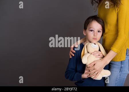 Sad boy with black eye, hurt bruised eye, hugging mom and teddy bear at home Stock Photo