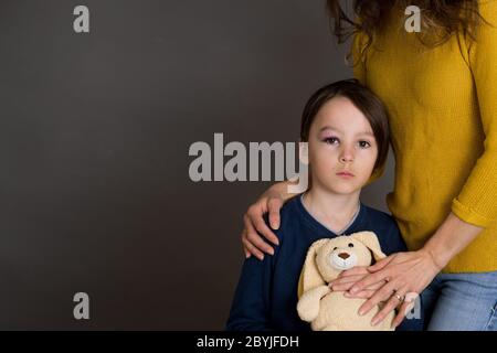 Sad boy with black eye, hurt bruised eye, hugging mom and teddy bear at home Stock Photo