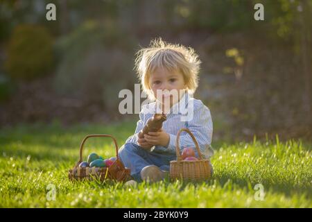 Little toddler boy, eating chocolate bunny in garden on sunset, easter eggs around him Stock Photo