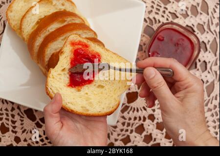Traditional Brazilian homemade bread - Hands putting strawberry jam on bread. Crochet towel background. Close-up. Top view. Stock Photo