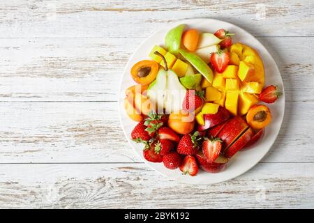 Healthy fruit platter of strawberries, mango, apples, pears, apricots, with peanut butter and peanuts on a white plate on a white wooden table, top vi Stock Photo
