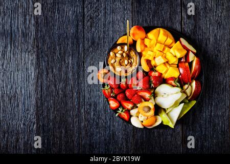 Healthy dessert of fresh fruits and berries with peanut dip: strawberries, mango, apples, pears, apricots and peanuts on a black plate on a dark woode Stock Photo