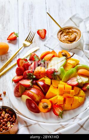 Fruit platter snack of fresh fruits and berries: strawberries, mango, apples, pears, apricots, with peanut butter and peanuts on a white plate on a wh Stock Photo