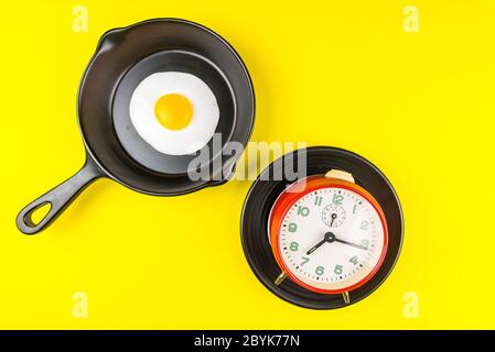 Frying pan with fried egg and alarm clock in a black plate isolated on a yellow background viewed from above.Concept of breakfast at the race against Stock Photo
