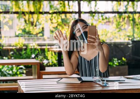 Happy Asian woman smiling wearing face shield holding a mobile phone on terrace. Video calling technology during Covid-19 - Corona Virus Stock Photo