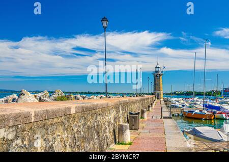 Stone pier mole with lighthouse, street lights and yachts on boat parking port marina in Desenzano del Garda town, blue sky white clouds background, Lombardy, Northern Italy Stock Photo