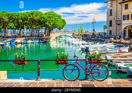 Bicycle bike near fence of old harbour Porto Vecchio with motor boats on turquoise water and Venetian bridge in historical centre of Desenzano del Garda town, blue sky, Lombardy, Northern Italy Stock Photo