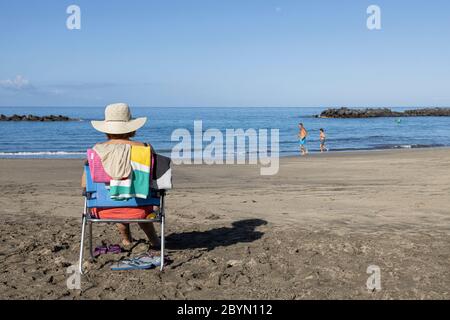 Woman in straw hat sitting on a portable chair on the Playa del Duque beach, Costa Adeje, Tenerife, Canary Islands, Spain Stock Photo