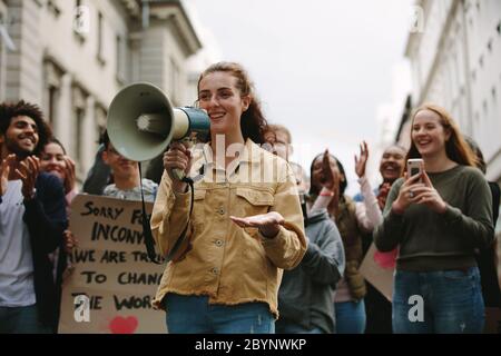 Woman with a megaphone in a rally outdoors on road. Woman standing outdoors with group of demonstrator clapping and celebrating. Stock Photo