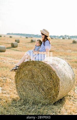 Young beautiful woman in striped dress and straw hat sits on hay bale and hugs her adorable little baby girl, enjoying summer sunset in field with Stock Photo