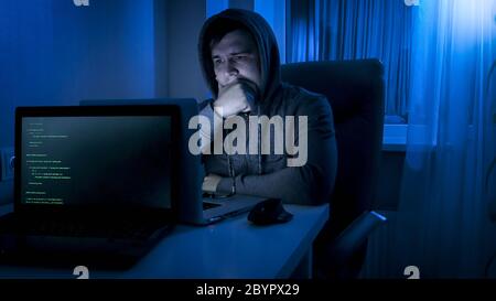 Thoughtful programmer in hood looking on laptop screen while working at night Stock Photo