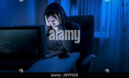 Angry male hacker in hood holding hands on his forehead while working at night Stock Photo