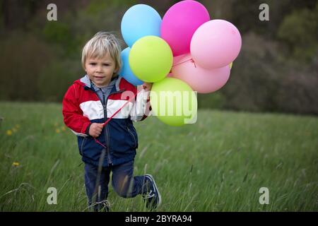 Little child, cute boy on a spring cold windy rainy day, holding colorful balloons in a field, running happily Stock Photo