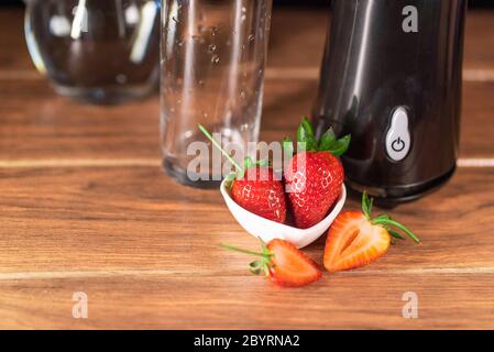 large red strawberries on a small white ceramic plate with a blender and a glass cup on a brown wooden table Stock Photo