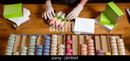 Female hands in gloves packs traditional french dessert macaroon in paper boxes for delivery. Take away of sweets concept Stock Photo