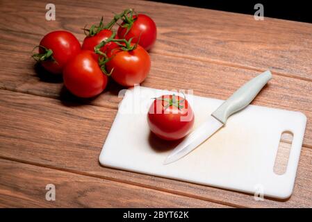 Fresh red tomato on white cutting board with knife next to it and many red tomatoes behind on brown wooden table Stock Photo