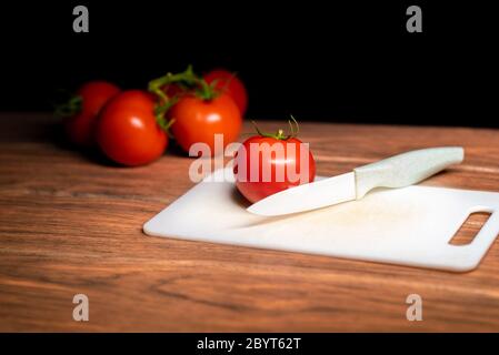 Ripe red tomato on a cutting board food and white ceramic knife with more red tomatoes in the background Stock Photo