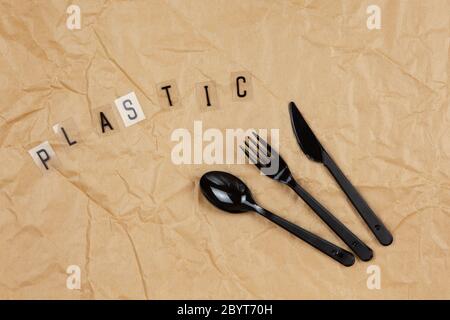 Disposable black appliances fork, spoon, knife, inscription Plastic from letters on transparent base on brown crumpled craft paper. Eco, zero waste co Stock Photo