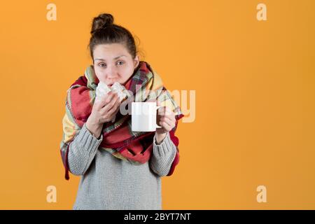 A young woman with a hot drink wrapped in a blanket wipes her nose with a napkin Stock Photo