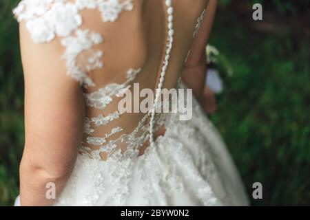 Bride in wedding dress. Close up of lace decorated back of white wedding gown and dense row of white buttons on spine. Outdoor background. Wedding day Stock Photo