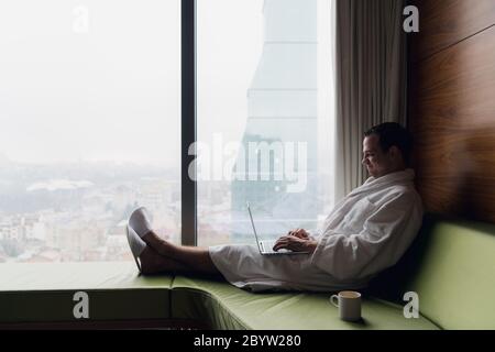 Young man is working on a laptop in his bed on a background of a panoramic window overlooking the skyscrapers. Freelancer, remote work, work from home Stock Photo