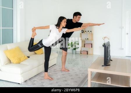 Young Asian couple doing yoga practice together and looking TV at home, man and woman working out together standing in living room, fit pair performin Stock Photo