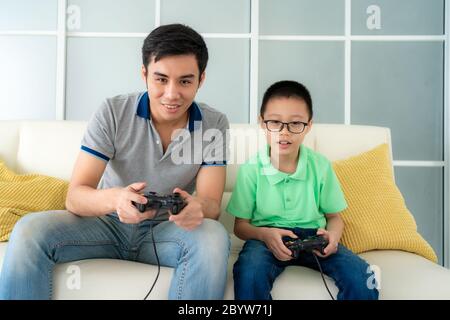 Asian family father and his son to playing video games with joysticks while sitting in sofa in living room at home, concept of family values and the m Stock Photo