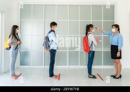 Three Asian student wearing mask standing distance of 6 feet from other people keep distance while teacher using thermometer temperature screening stu Stock Photo