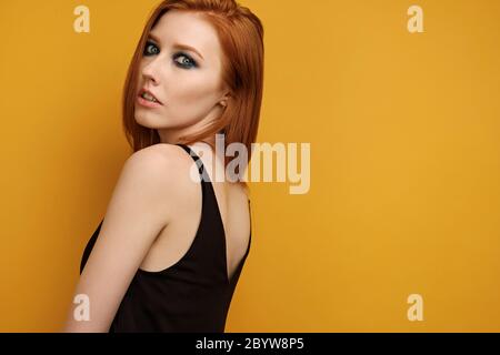 Red-haired girl in a black top and with blue eye make-up stands on a yellow background, looking into the frame over her shoulder Stock Photo
