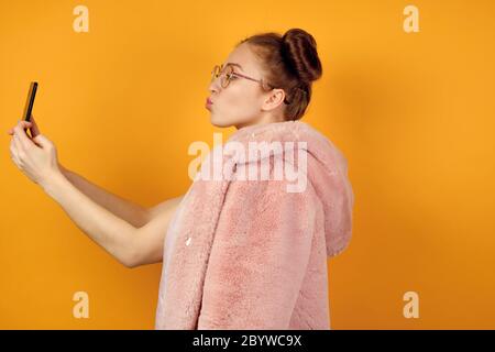 A red-haired girl in round glasses stands in profile in a pink coat and takes a selfie, stretching out lips in a kiss Stock Photo