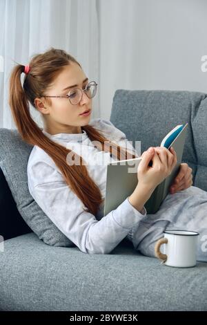 Redhead girl in pajamas and round glasses lies on the sofa and reads a book. Stock Photo