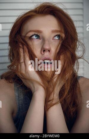 Headshot. The red-haired girl looks up, opening her mouth and pressing her hands to her face. Stock Photo