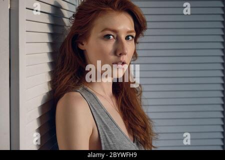 A red-haired girl stands in a half-turn against a white screen and looks into the frame, in the rays of the setting sun. Stock Photo