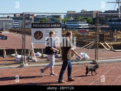 Baltimore, Maryland, USA. 8th June, 2020. Side view of woman with face mask around her wrist walking small dog on leash, with man beside her wearing mask on his face, a trio of long shadows cast behind them, and boats on the water in the background. They pass in front of the construction site of the Inner Harbor Water Taxi Terminal that has a partially obscured sign on the surrounding chain-link fence reading 'Progress Ahead', detailing the City of Baltimore Department of Transportation project. Kay Howell/Alamy Stock Photo