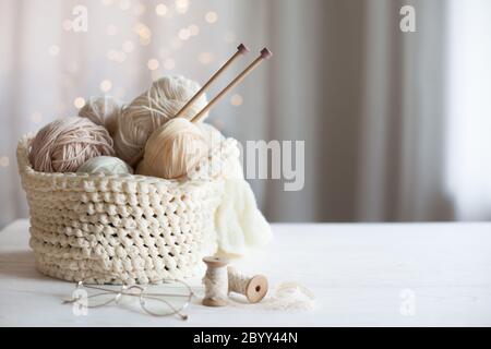 Knitted white and beige fabric. Woolen and cotton yarn lies in a basket. Glasses lie on a white wooden background. Stock Photo