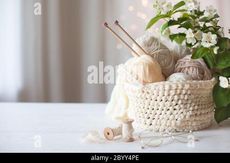 Knitted white and beige fabric. Woolen and cotton yarn lies in a basket. Glasses lie on a white wooden background. Stock Photo
