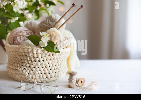 Knitted white and beige fabric. Woolen and cotton yarn lies in a basket. Glasses lie on a white wooden background. Stock Photo