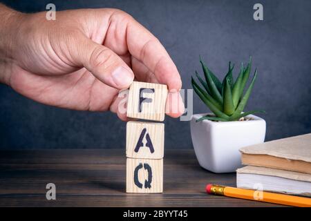 FAQ FREQUENTLY ASKED QUESTIONS. Wooden blocks and a man's hand. Books, pencil and green plant on a wooden table Stock Photo