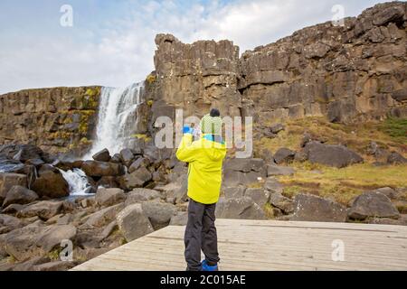 Cute child, boy, enjoying a sunny day in Oxararfoss Waterfall in Thingvellir National Park rift valley, taking pictures with cellphone, Iceland autumn Stock Photo