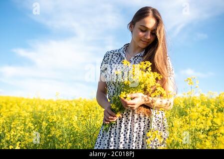 Beautiful girl in a rapeseed field, with a bouquet in her hands against a blue sky. Stock Photo