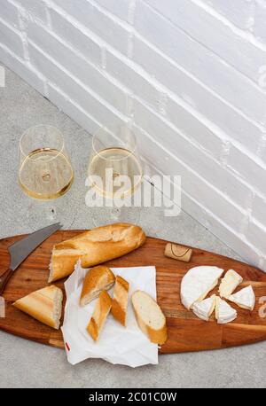 Slices of camembert cheese, baguette and two glasses of white wine on wooden board. White brick wall as background. Stock Photo