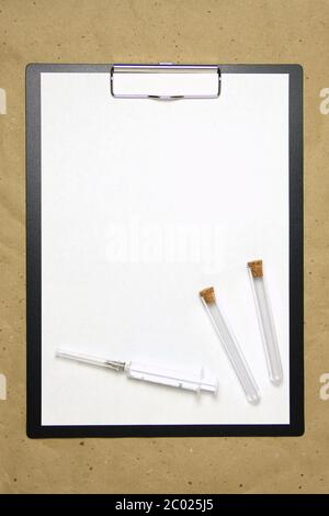 The tablet with a clip for the clip of paper with a white sheet a4 lies against the background of craft brown crumpled paper. On top is a medical syringe and two transparent empty tubes with stoppers. Place for text and layout for design. Stock Photo
