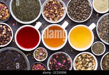 Assortment of tea cups, dried leaves, fruit and herbs in bowls on dark, stone background, top view Stock Photo