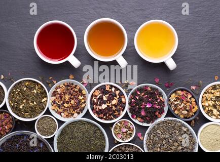 Assortment of tea cups, dried leaves, fruit and herbs in bowls on dark, stone background, top view Stock Photo